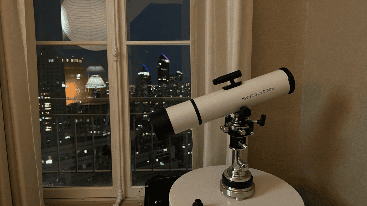 A compact telescope on a table beside an apartment window at night with city lights outside