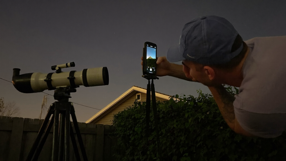 A beginner in a suburban backyard using a smartphone mounted near a telescope on an alt-azimuth tripod at night