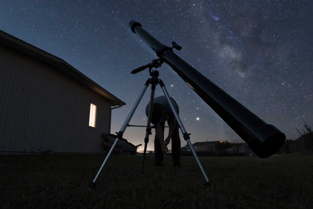 A refractor telescope on a tripod angled up at the Milky Way in a suburban backyard, an observer bending to the eyepiece, one lit house window behind