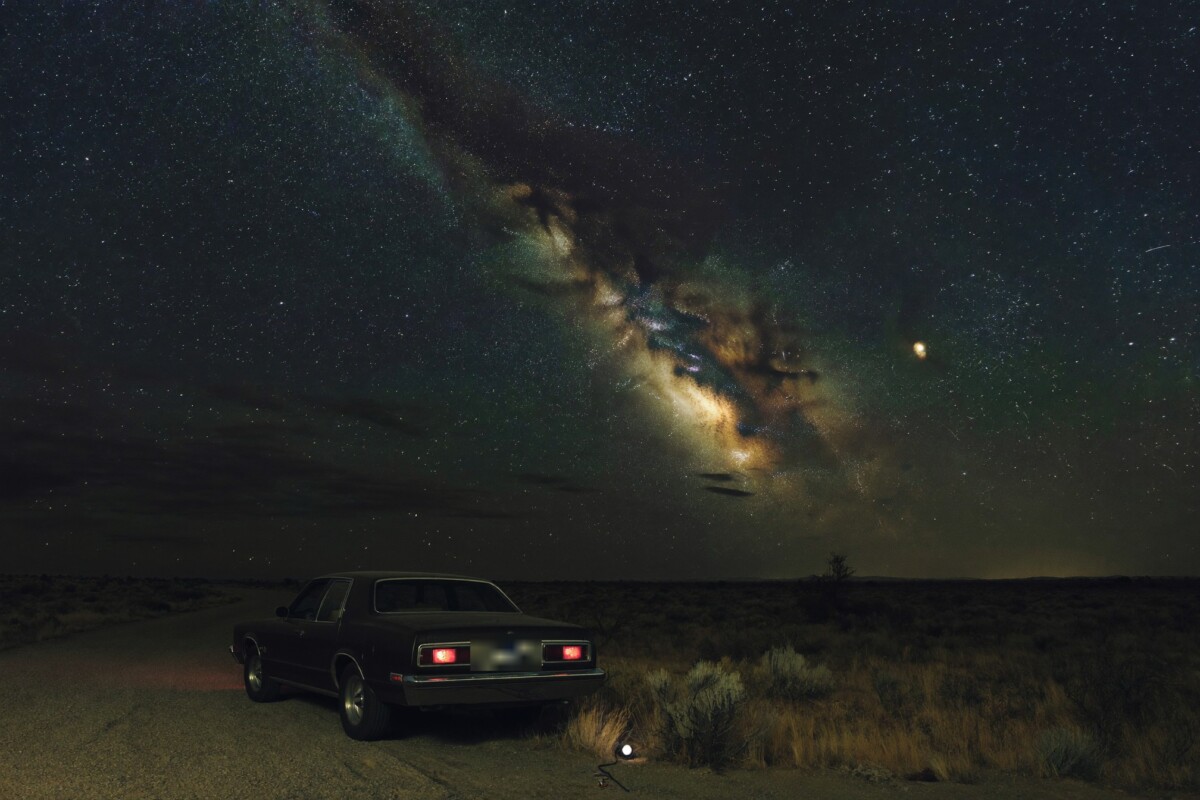 A car parked on a remote gravel pullout beneath the arch of the Milky Way in open desert scrub