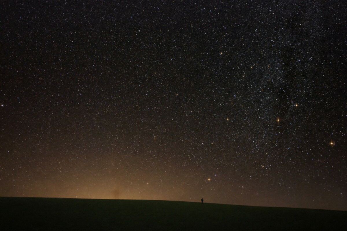 A dense field of stars above a low rural horizon with a faint orange light dome from a distant town